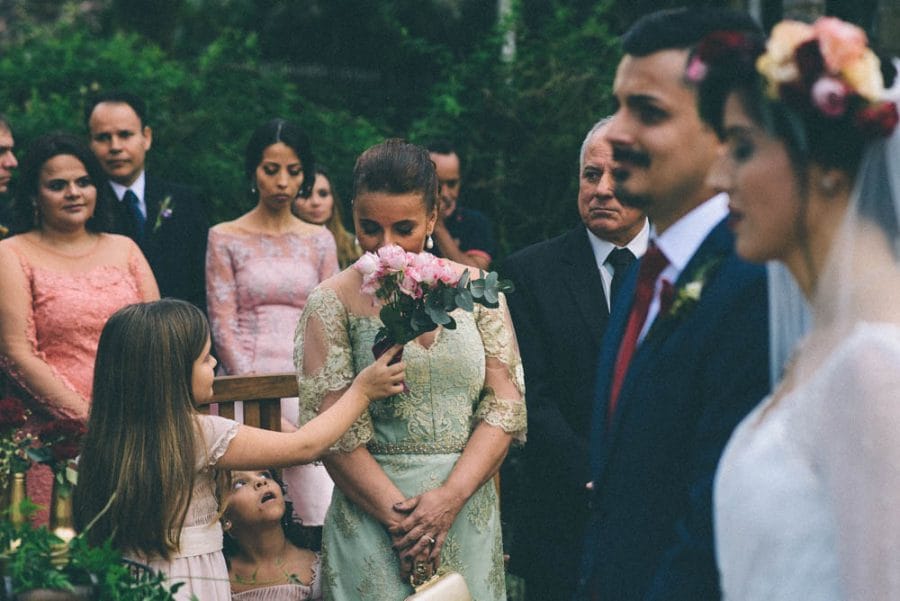 Wedding photography photo during a ceremony with the bride and groom to the right of the frame slightly out of focus and with a flower girl telling her mom or aunt to smell the flowers