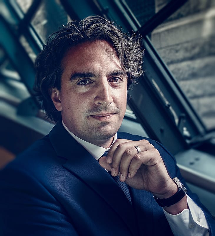 Image of a corporate portrait of a man with medium-length curly hair in a blue suit, a watch on, a ring in front of a window being lit with window light