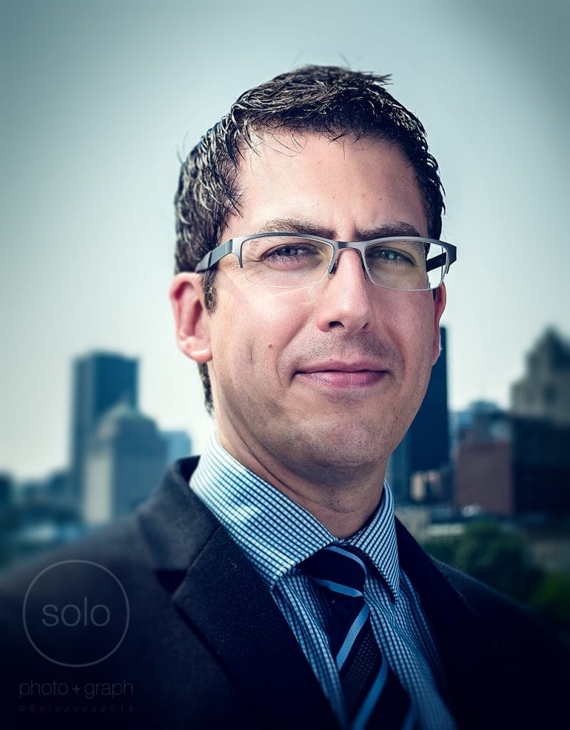 Image of a corporate employee in a suit and glasses straight on looking at the camera in front of a cityscape of buildings