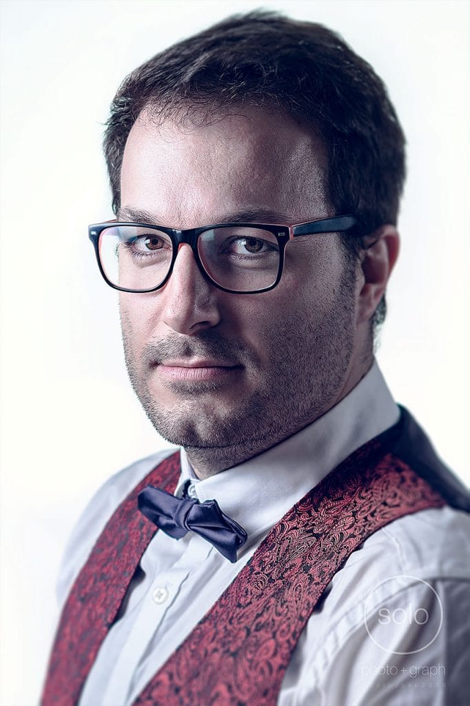 Photo of a corporate portrait lit with a pure white background of a man with brown hair, glasses, a bowtie, and a red vest