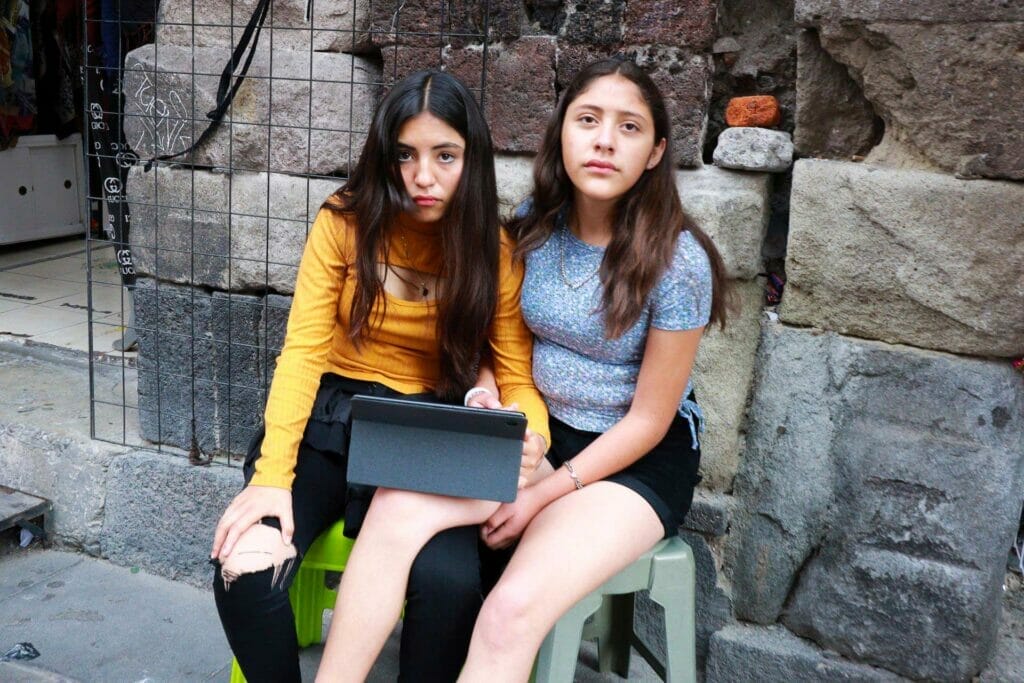 Two girls sitting on plastic chairs using a tablet on a sidewalk