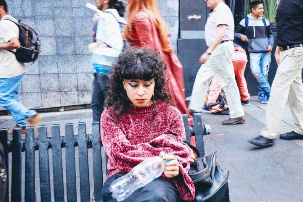 A woman looking down after taking a drink from a plastic water bottle while sitting on a bench on the street