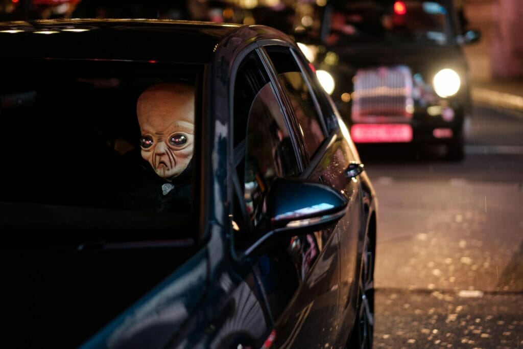 A person in an alien mask in a dark car in front of a British taxi.