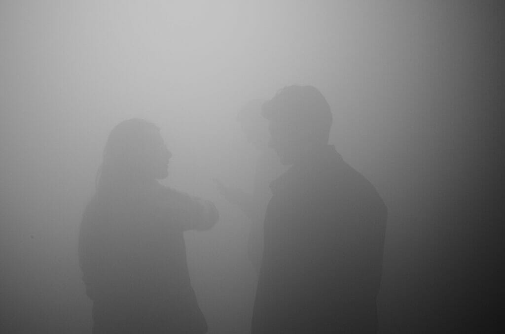 Silhouettes of a man and woman talking in gray fog and a silhouette behind them of someone on their phone.