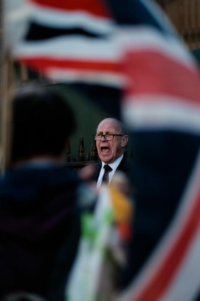 An old man yelling with black glasses, and a black suit and tie.