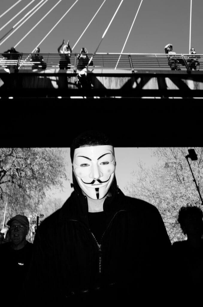 A person wearing a Guy Fawkes mask under a bridge.