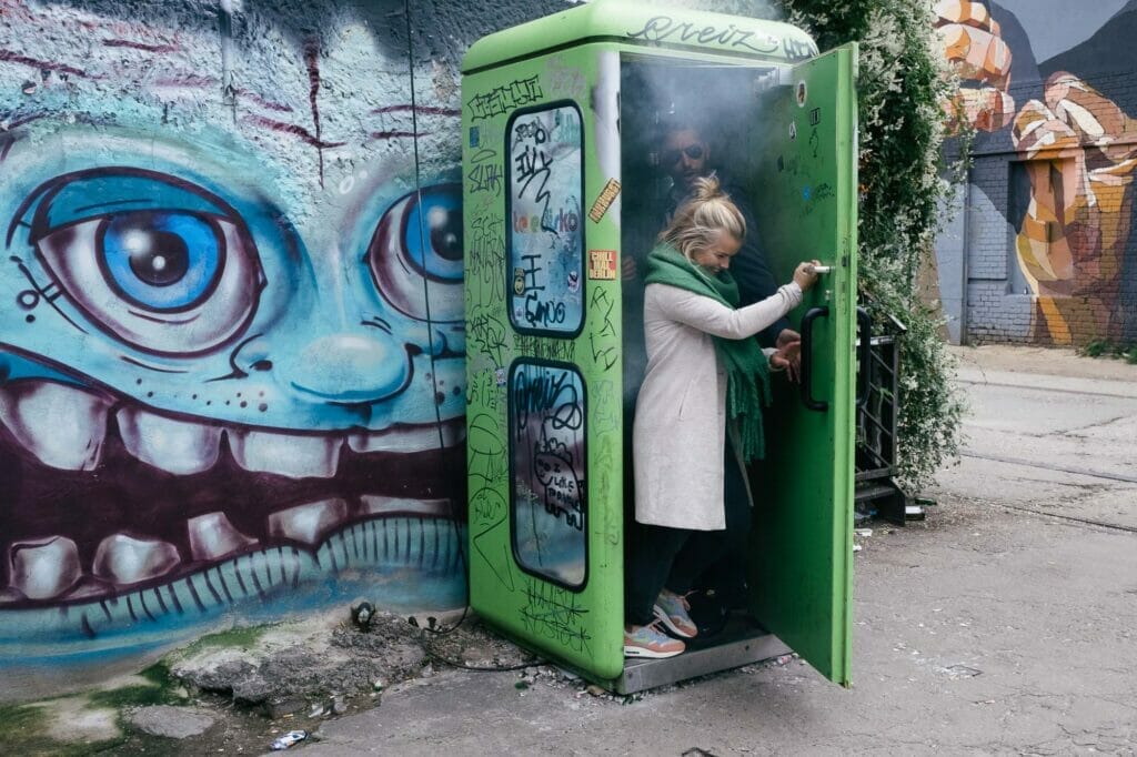 A blonde woman exiting a green box on the street with smoke coming out of it.