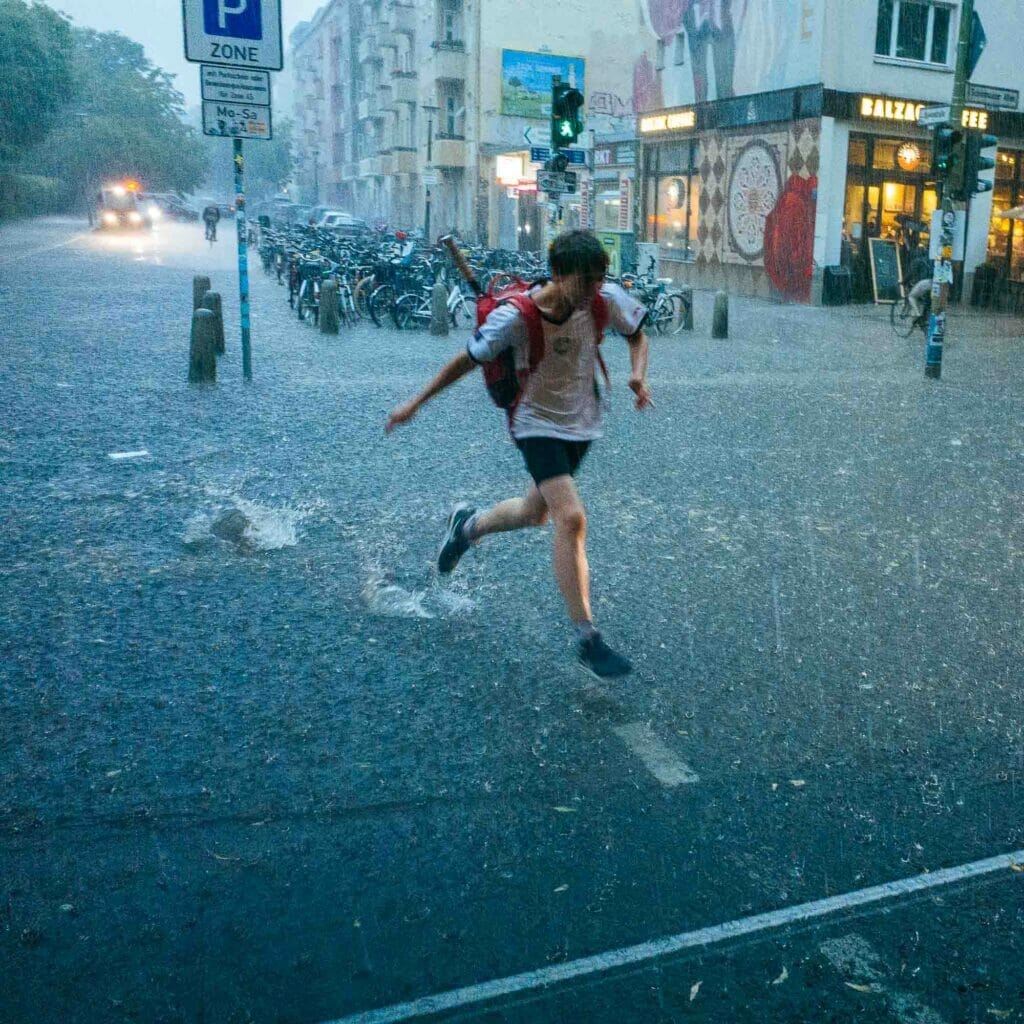 A boy carrying a tennis racquet in a red backpack running in a rainstorm.