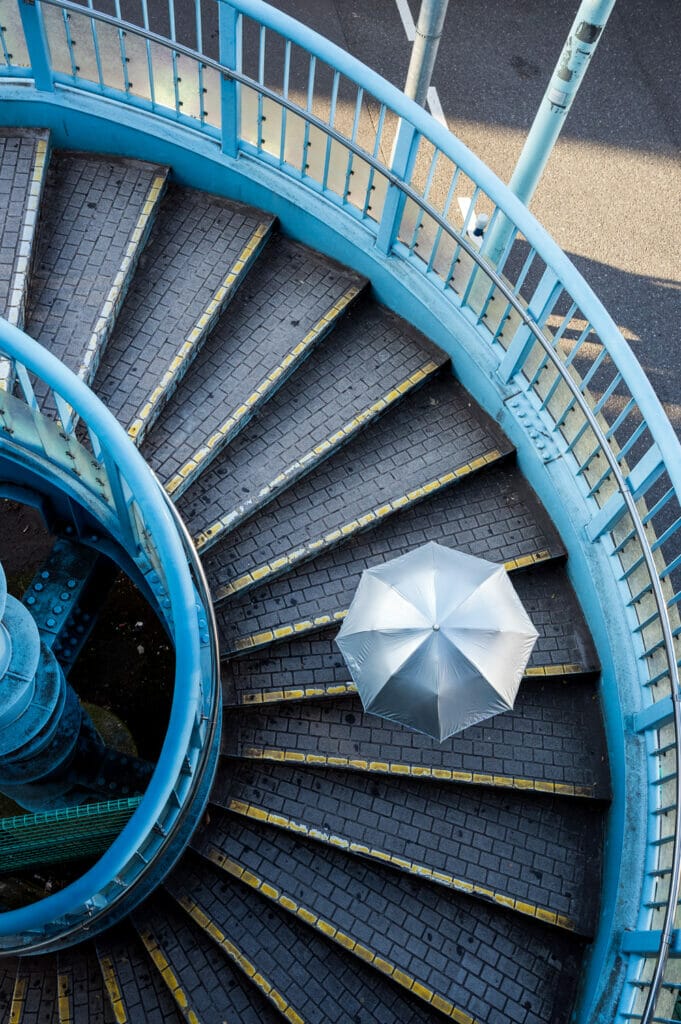 Looking down at a spiral staircase as a person walks down the stairs. You can only see the silver umbrella and not the person below it.